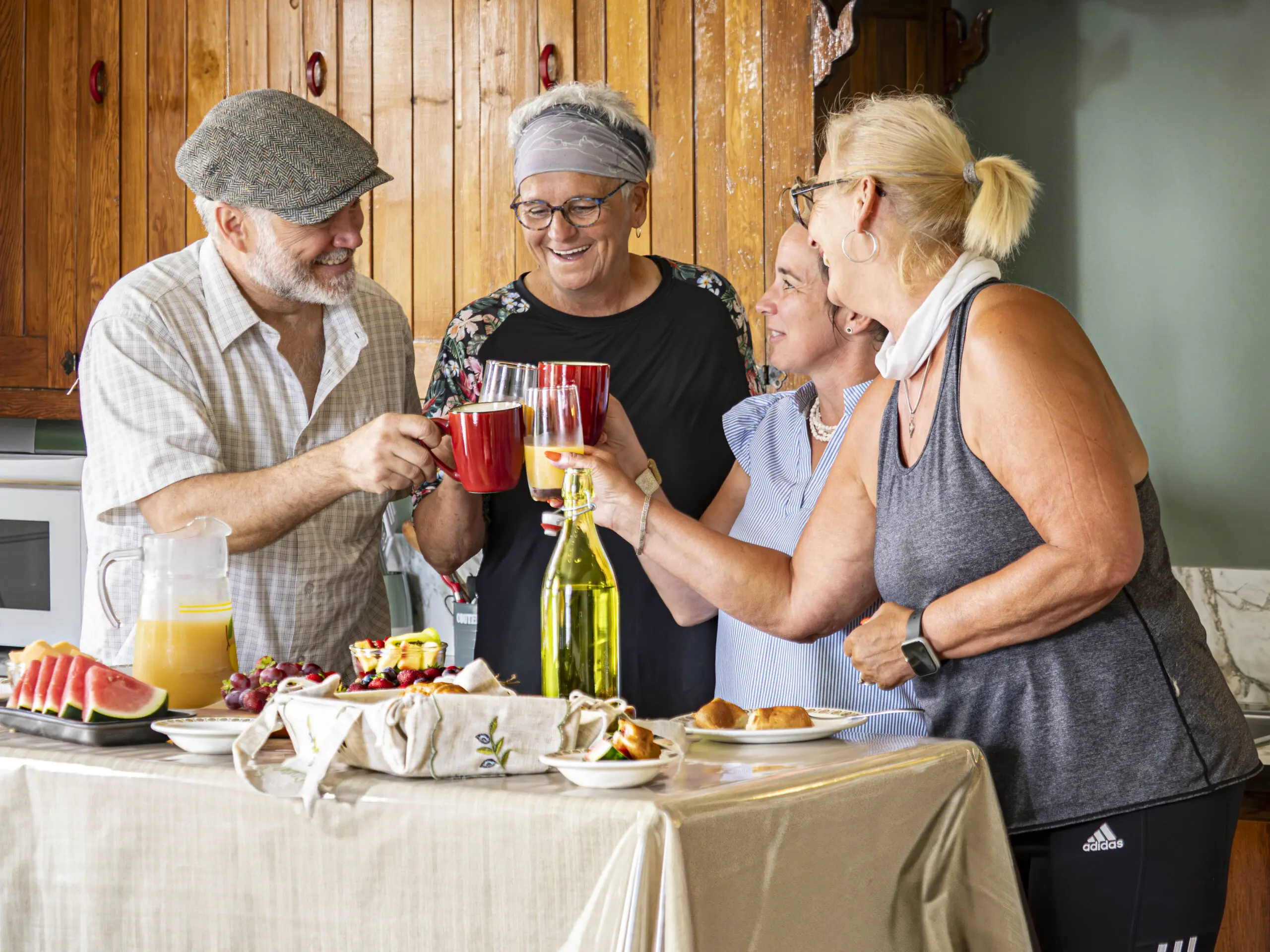 Quatre personnes âgées se tiennent autour d'une table garnie de nourriture et de boissons, souriant et trinquant avec des verres. La table contient des pastèques, des raisins, des pâtisseries et des bouteilles, et ils semblent profiter d'une joyeuse réunion à l'intérieur.