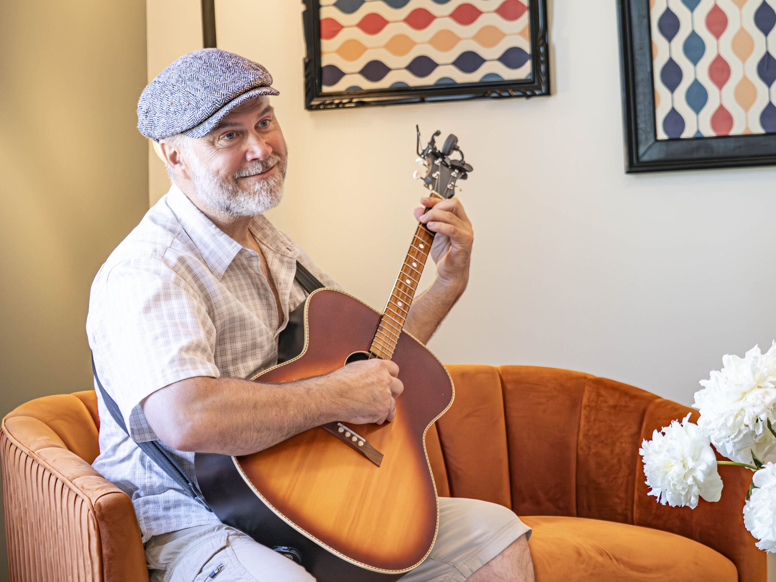 Un homme souriant à la barbe grise est assis sur une chaise orange et joue de la guitare acoustique. Il porte une casquette plate et une chemise à carreaux. Des œuvres d'art abstraites et des fleurs blanches sont visibles à l'arrière-plan.