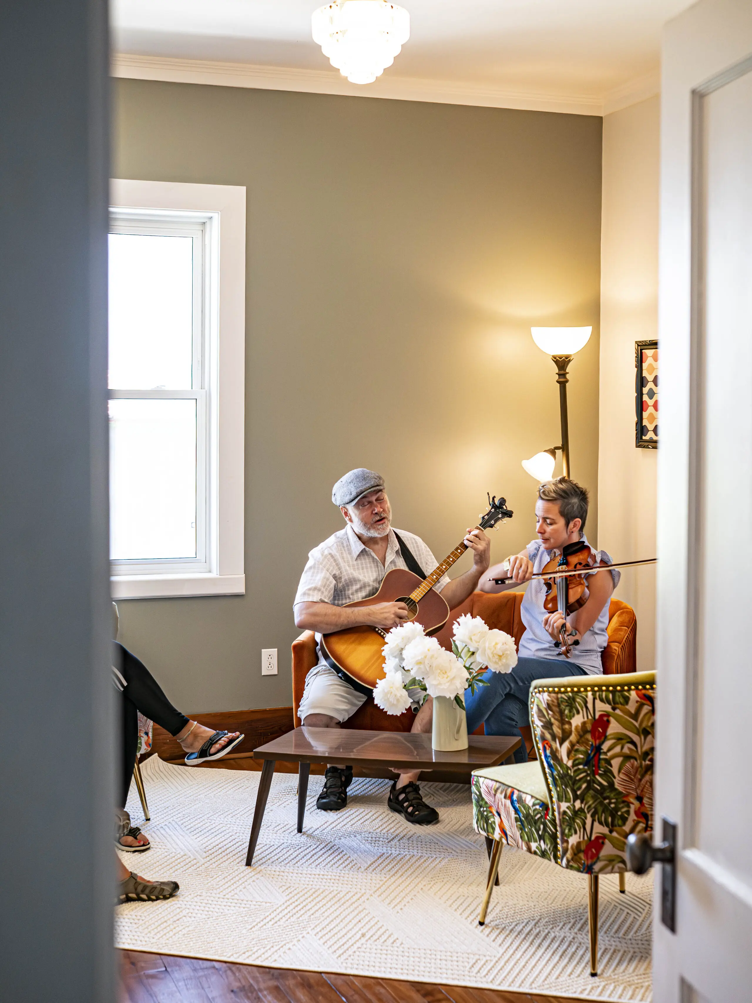 Deux personnes sont assises dans un salon confortable et jouent d'instruments de musique, l'une avec une guitare et l'autre avec un violon. Une chaise à fleurs et des fleurs blanches se trouvent au premier plan, la lumière naturelle entrant par une fenêtre.