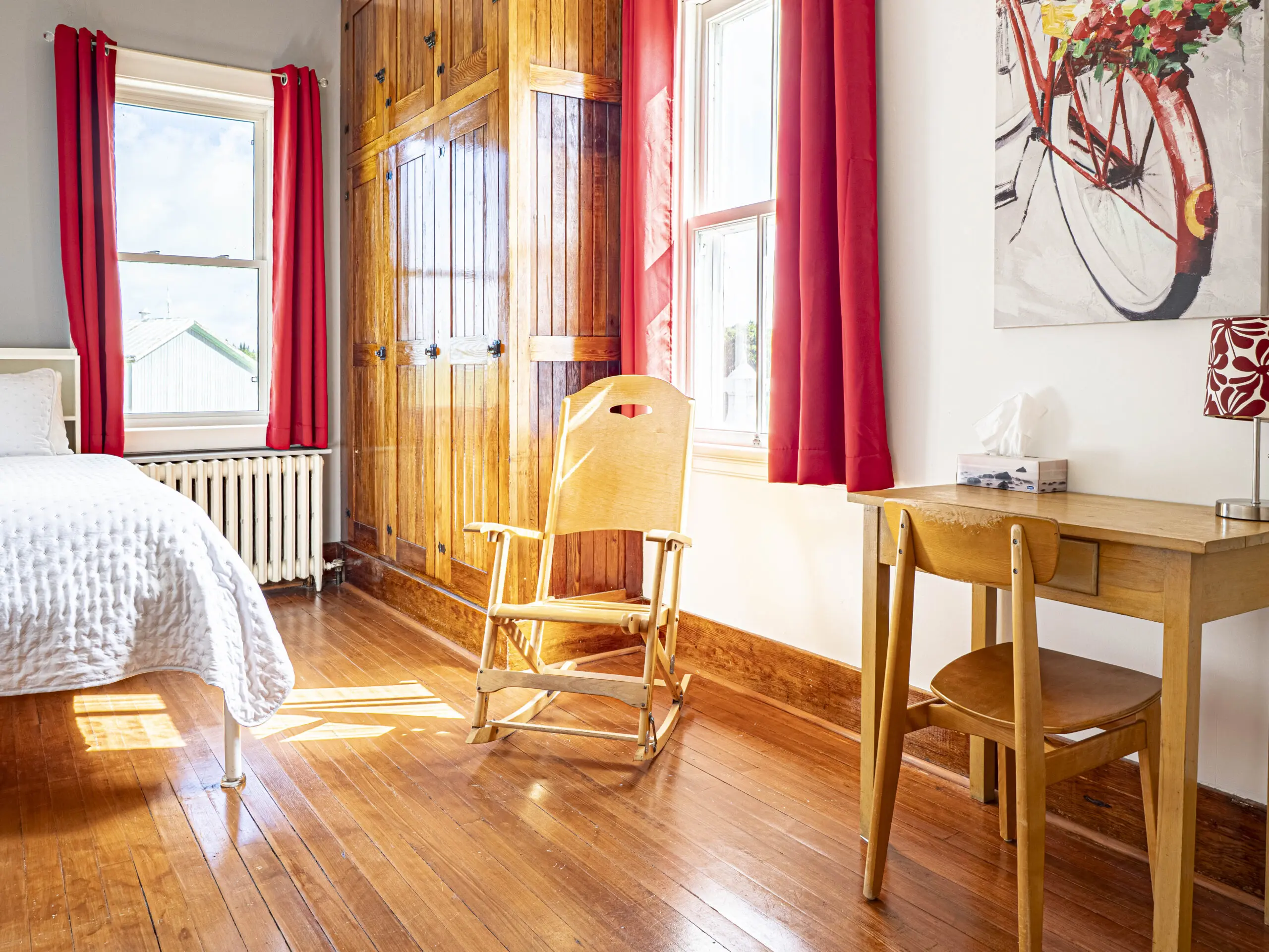 Une chambre lumineuse avec un sol en bois, un lit avec des draps blancs, une chaise à bascule en bois, un petit bureau avec une boîte à mouchoirs et une lampe, des rideaux rouges et une fenêtre qui laisse entrer la lumière naturelle.