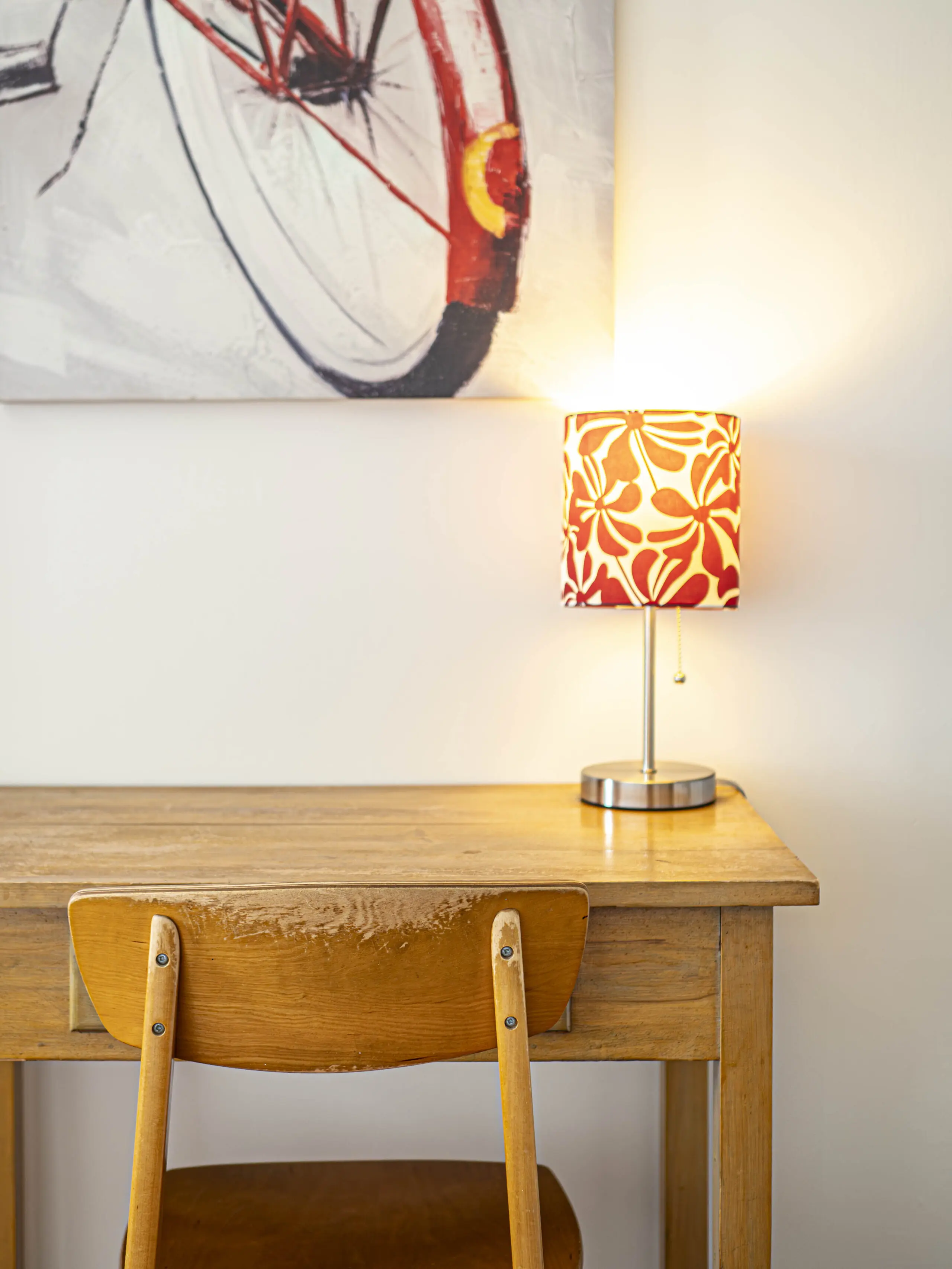 Un bureau et une chaise en bois sous un tableau mural représentant une bicyclette ; une lampe allumée avec un abat-jour floral rouge et blanc est posée sur le bureau, jetant une lueur chaleureuse sur le mur clair.