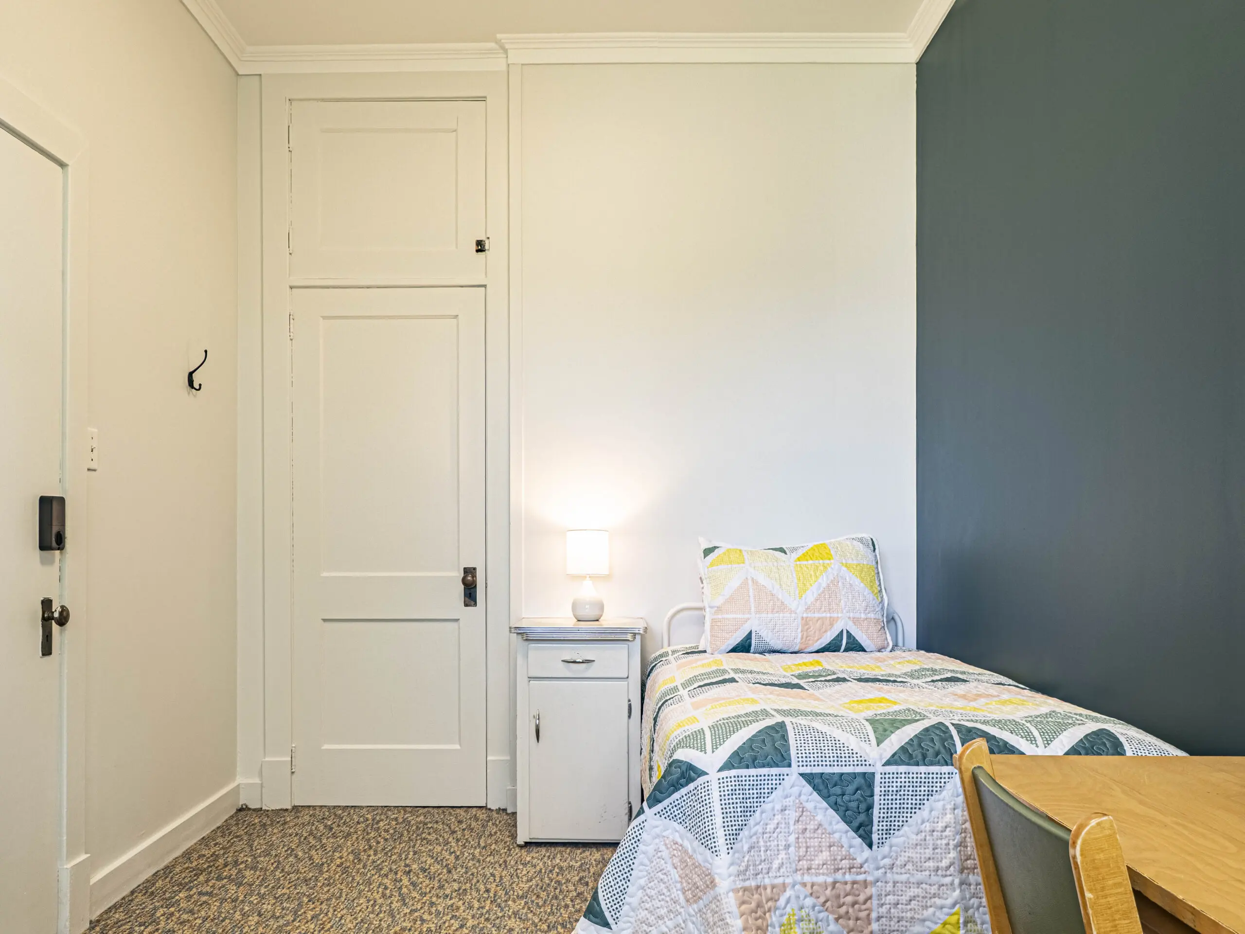 Un lit simple bien fait avec un édredon géométrique coloré est posé à côté d'une table de nuit blanche avec une petite lampe. Un bureau et une chaise se trouvent à proximité ; la pièce a des murs blancs, un mur d'accent sombre et une porte fermée.
