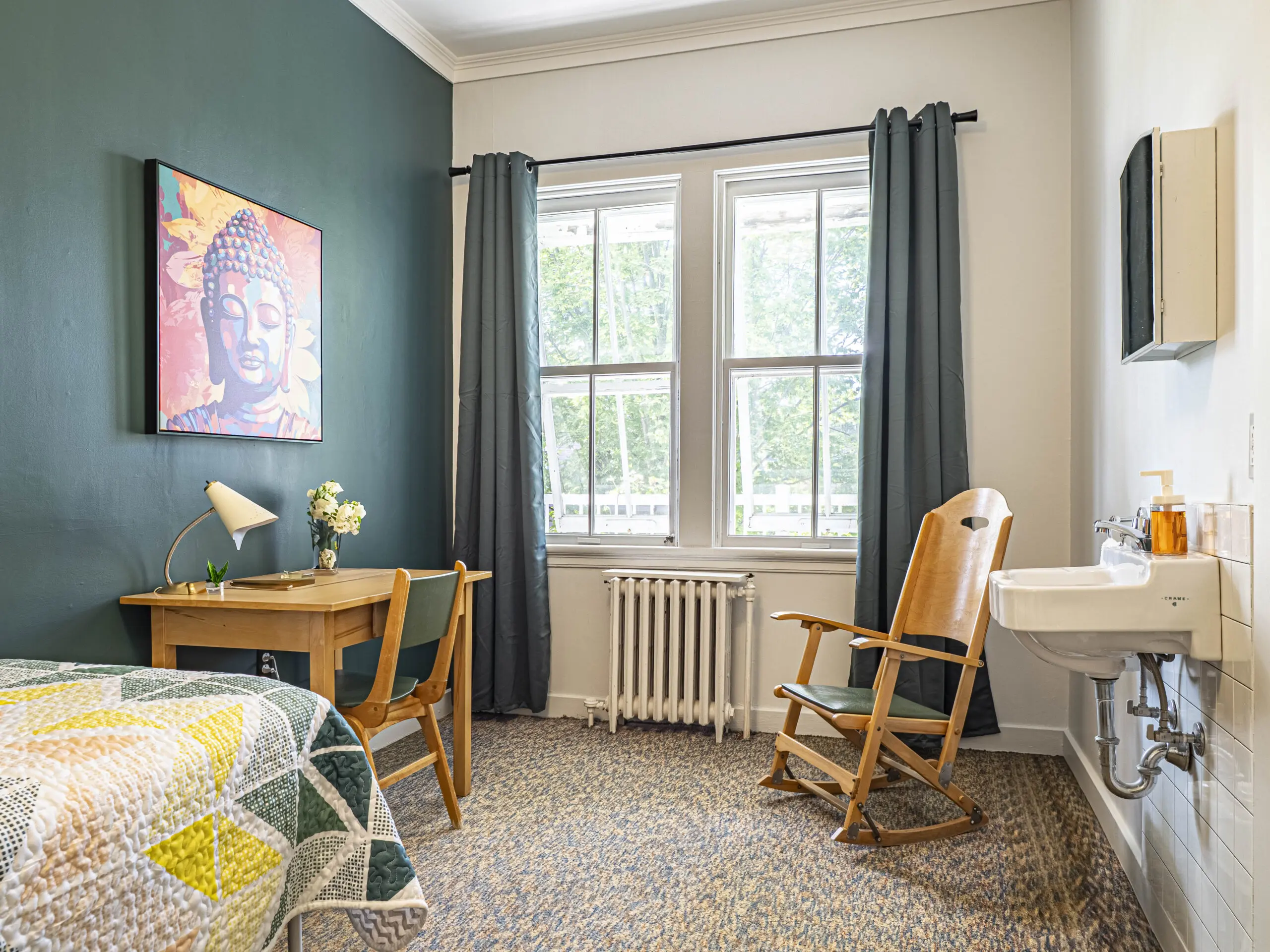 Une chambre confortable, éclairée par le soleil, avec un mur d'accent vert orné d'une œuvre de Bouddha, un bureau et une chaise en bois, un lit avec un édredon à motifs, un fauteuil à bascule, une fenêtre avec des rideaux sombres et un lavabo mural avec des articles de toilette.