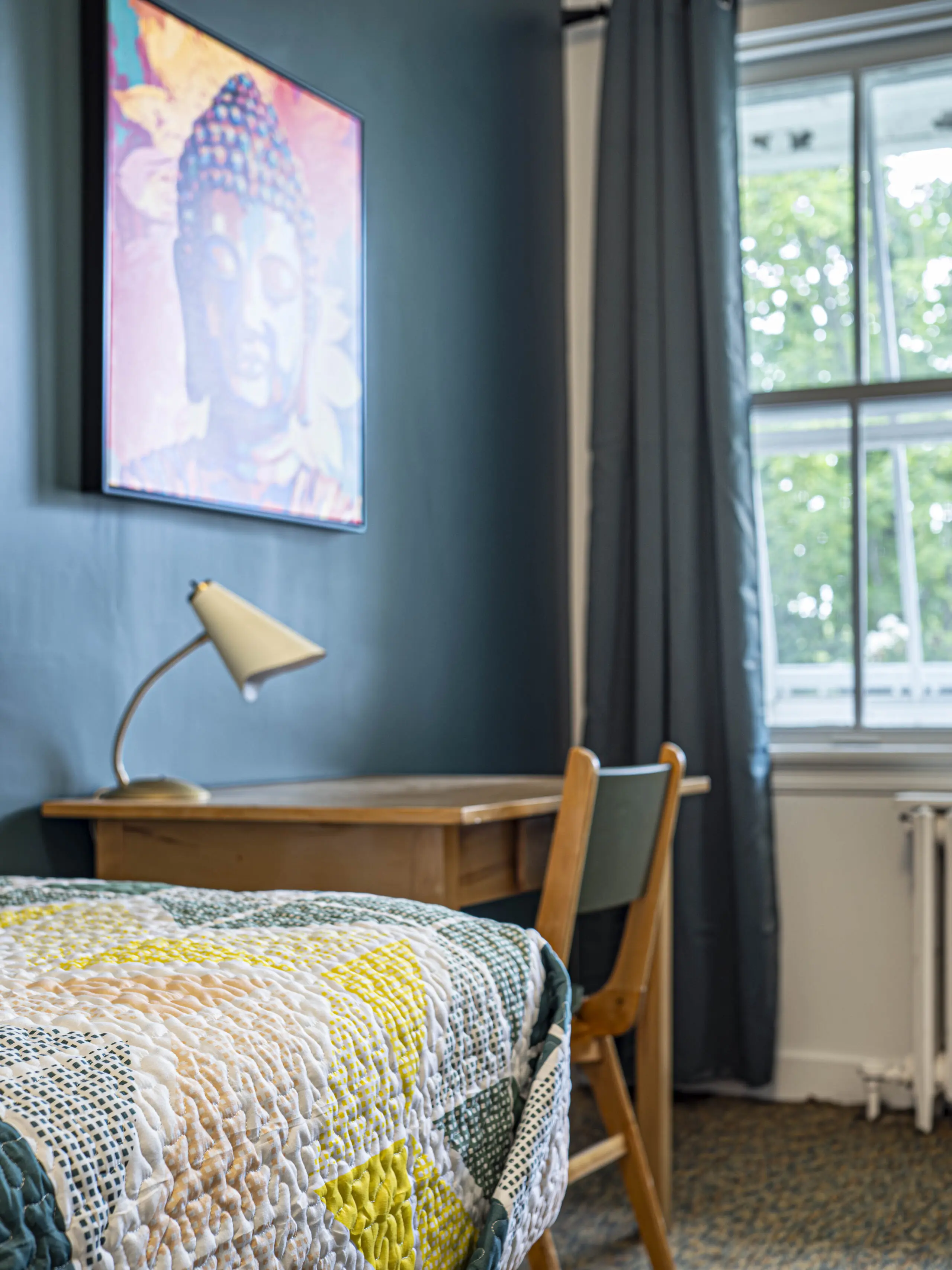 Un coin de chambre confortable avec un bureau et une chaise en bois près d'une fenêtre, une lampe sur le bureau, un lit avec un édredon coloré et une œuvre d'art de Bouddha accrochée à un mur sarcelle. Des arbres verts sont visibles à l'extérieur de la fenêtre.