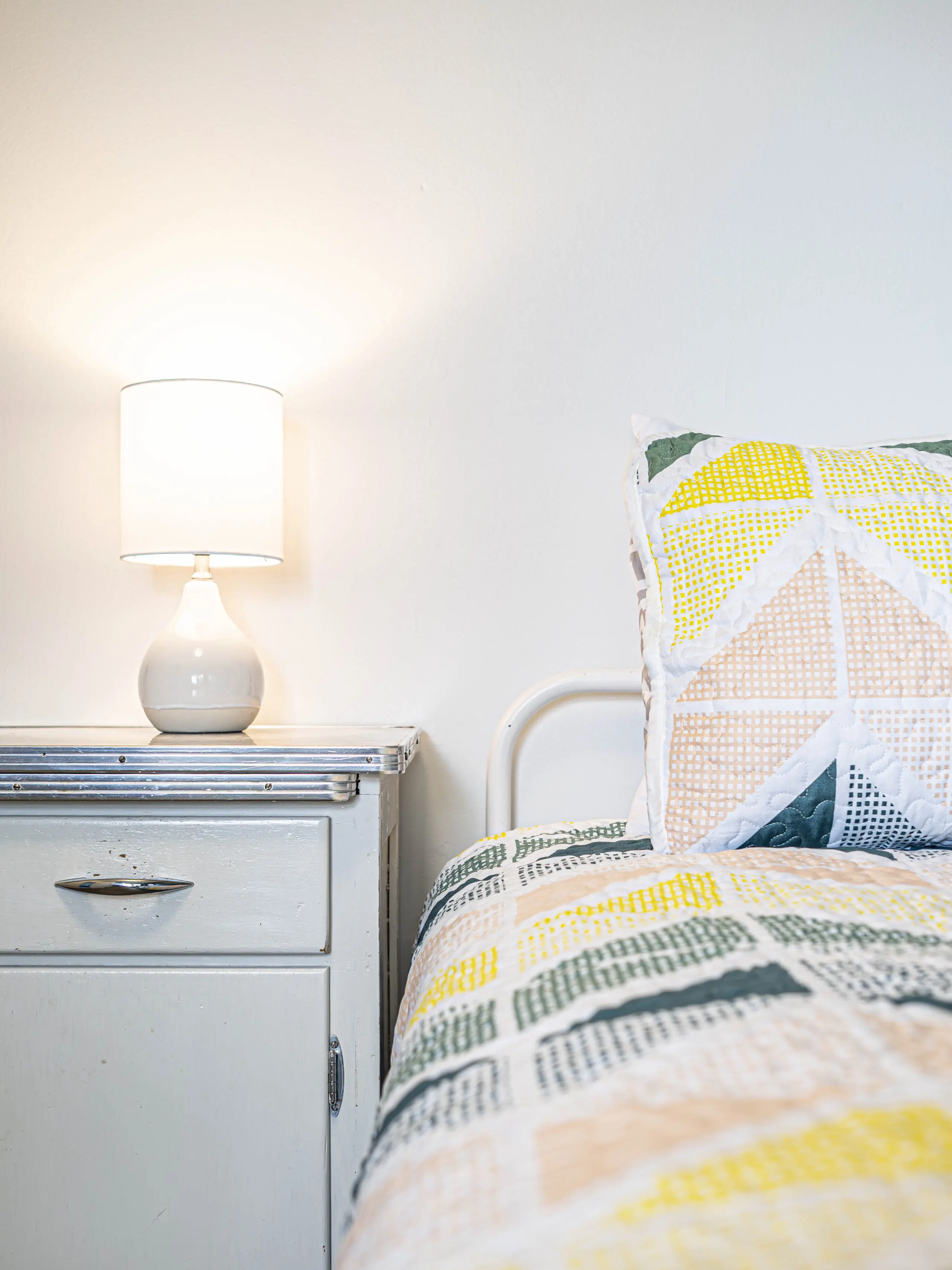 Une table de chevet avec une lampe blanche se trouve à côté d'un lit avec un édredon et un oreiller à motifs géométriques colorés. La scène est doucement éclairée, avec une esthétique propre et minimale.