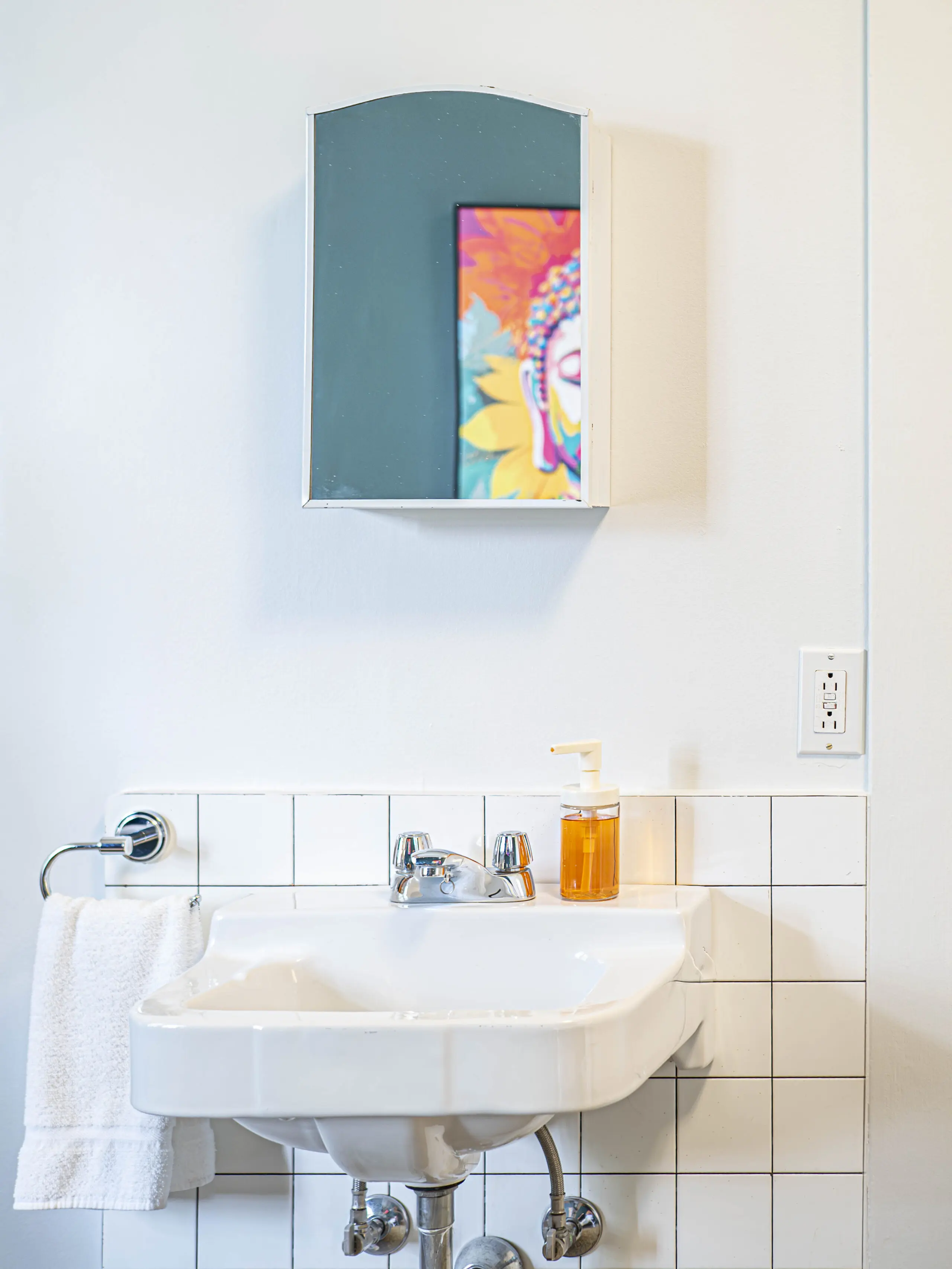 Un lavabo de salle de bains carrelé blanc avec une serviette suspendue à gauche, un distributeur de savon orange et une armoire à glace au-dessus. Une œuvre d'art colorée se reflète partiellement dans le miroir.