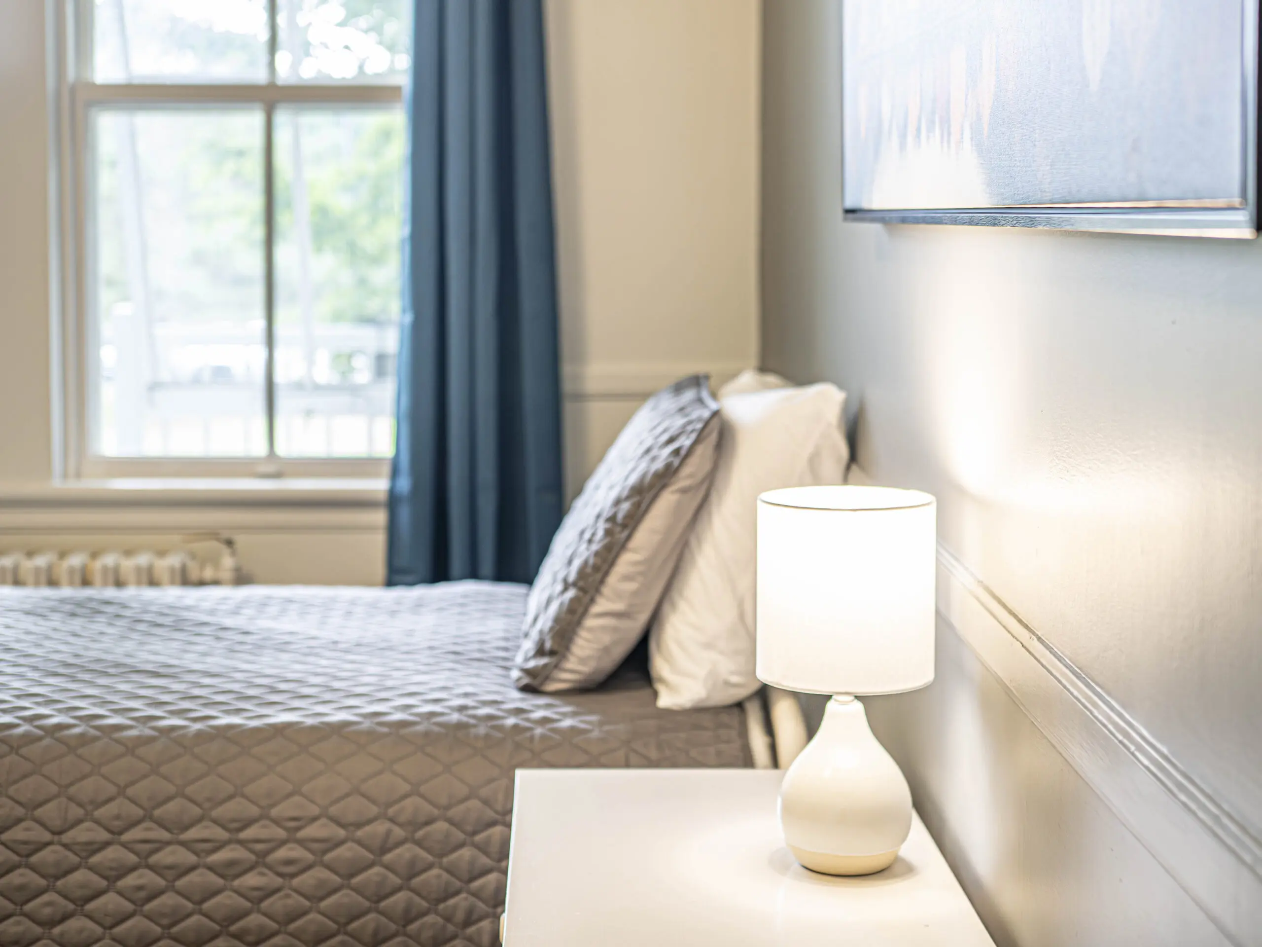 Un lit avec des draps et des oreillers gris matelassés se trouve à côté d'une table de nuit blanche avec une lampe blanche allumée. Une fenêtre aux rideaux bleus laisse entrer la lumière du jour, illuminant les murs peints avec douceur.