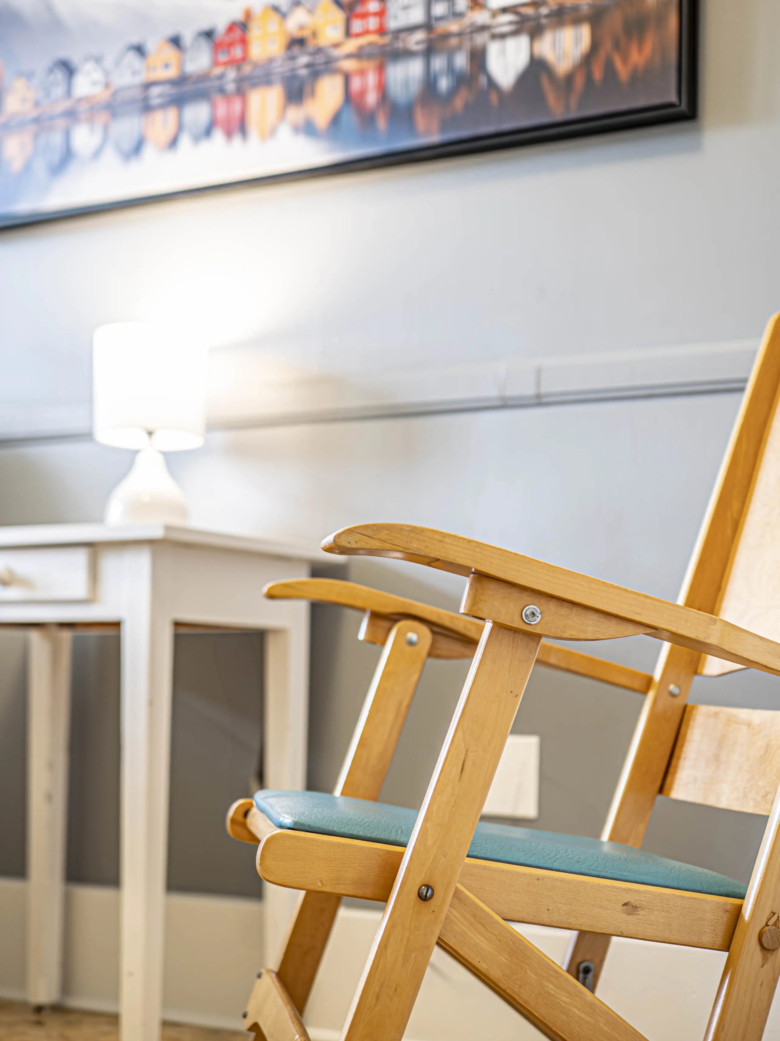Une chaise en bois avec un coussin sarcelle est assise près d'un bureau blanc et d'une lampe allumée ; une peinture colorée de maisons est accrochée au mur au-dessus de la scène.
