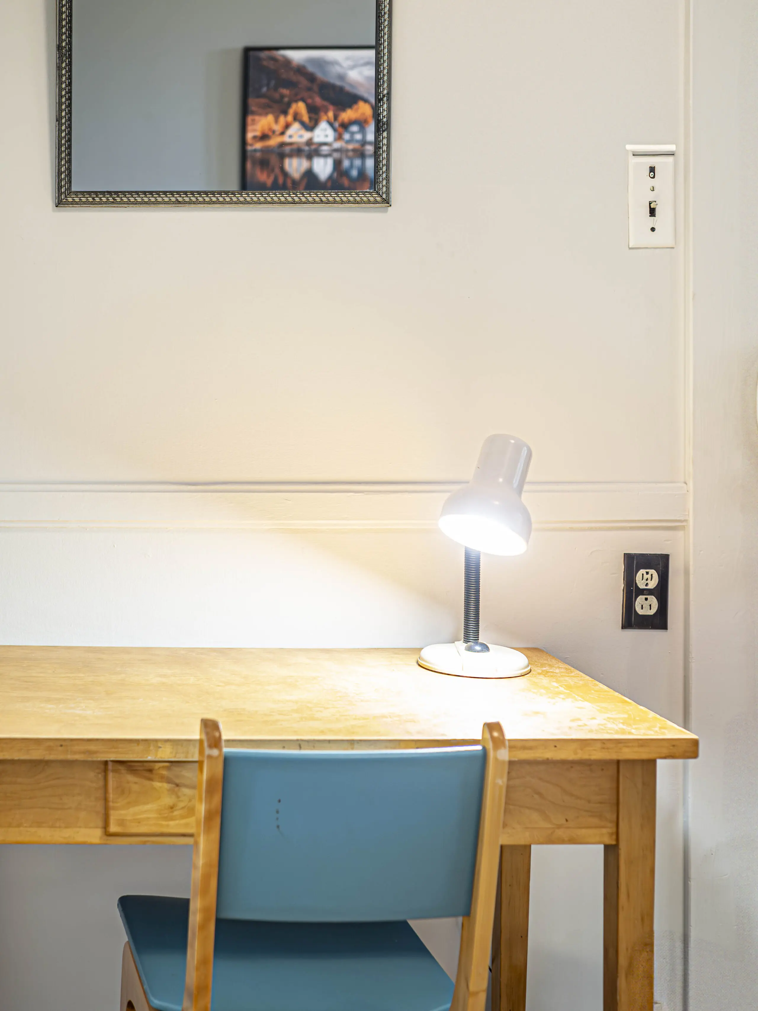 Un petit bureau en bois avec une chaise bleue est posé contre un mur blanc. Une lampe de bureau allumée est posée sur le bureau. Au-dessus, un miroir et une photo encadrée reflètent la lumière. Des prises électriques sont visibles sur le mur près du bureau.