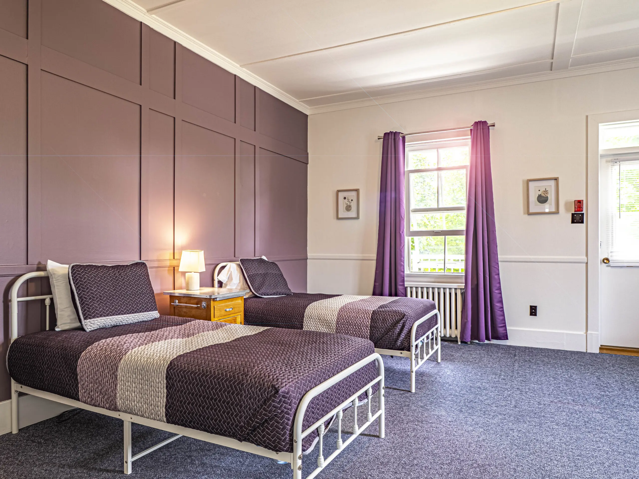 Une chambre lumineuse avec deux lits simples recouverts d'une literie violette, une table de nuit avec une lampe entre les deux, des rideaux violets sur une fenêtre, et des murs à panneaux violets au-dessus d'un lambris blanc.