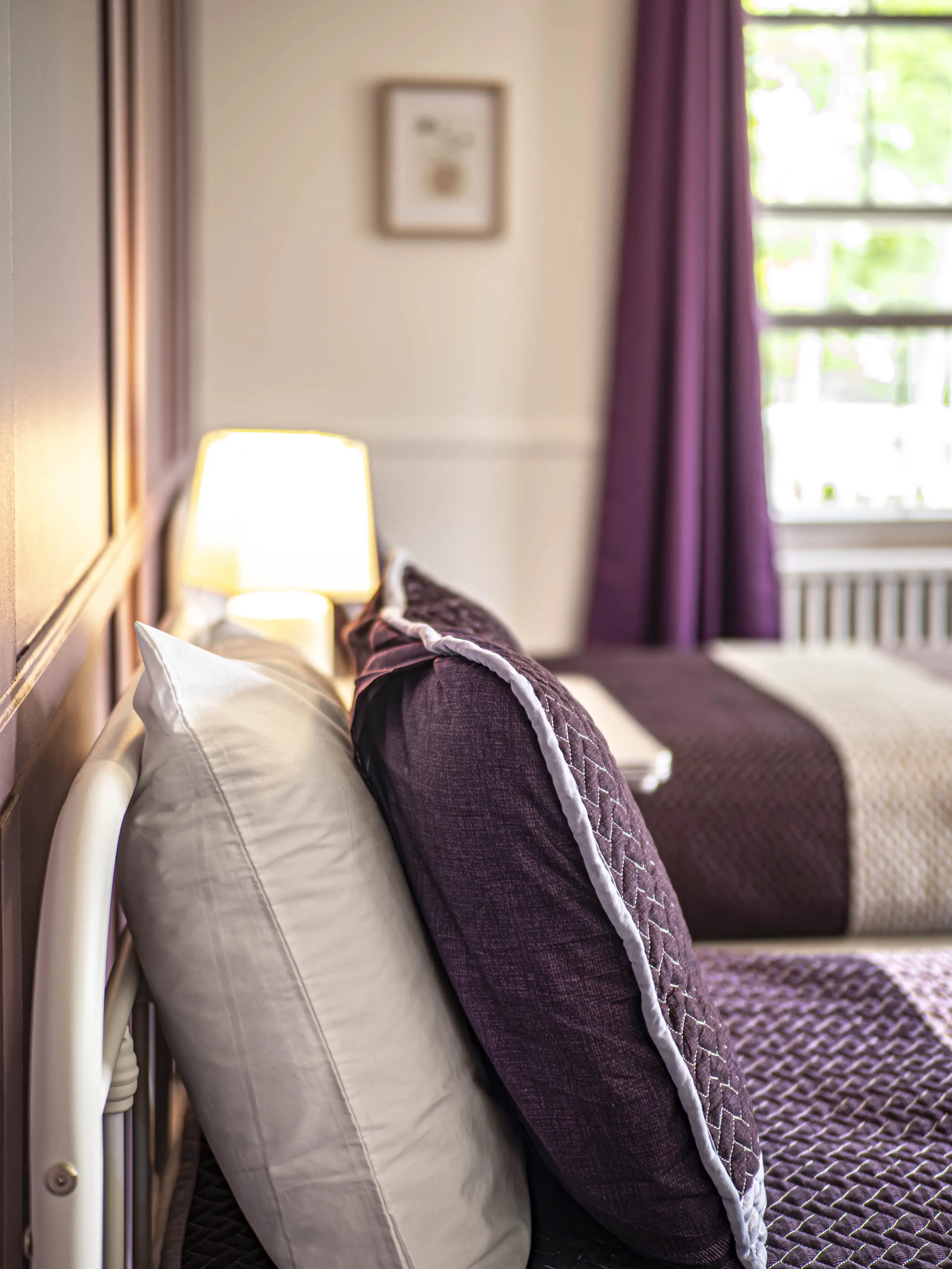 Gros plan sur un lit soigneusement fait avec des oreillers et des draps violets et beiges, une lampe allumée sur une table de chevet et une fenêtre avec des rideaux violets dans une chambre à coucher doucement éclairée.