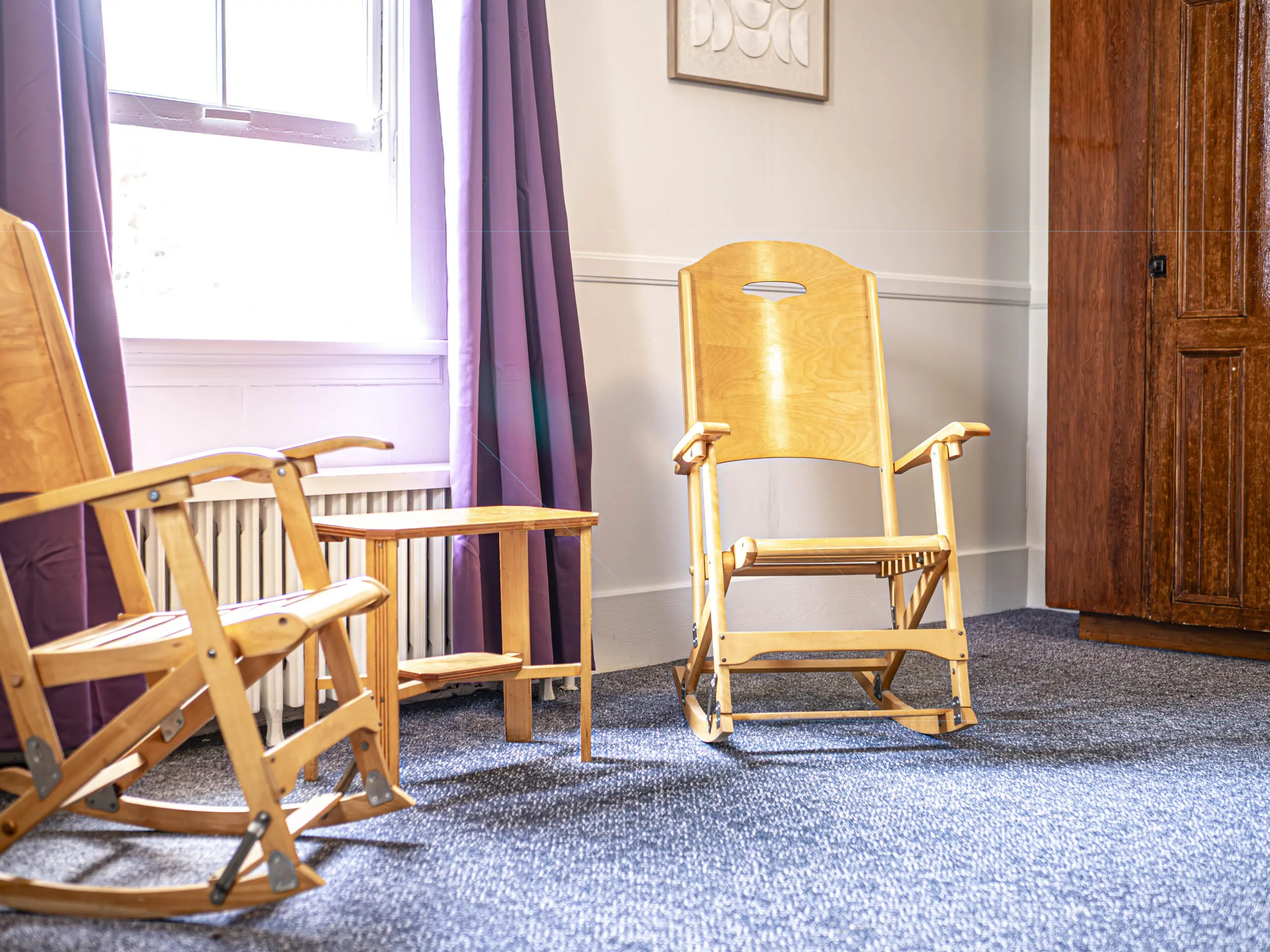 Deux fauteuils à bascule en bois et une petite table sont posés sur un tapis bleu dans une pièce ensoleillée avec une fenêtre, des rideaux violets, des murs clairs et une armoire en bois.