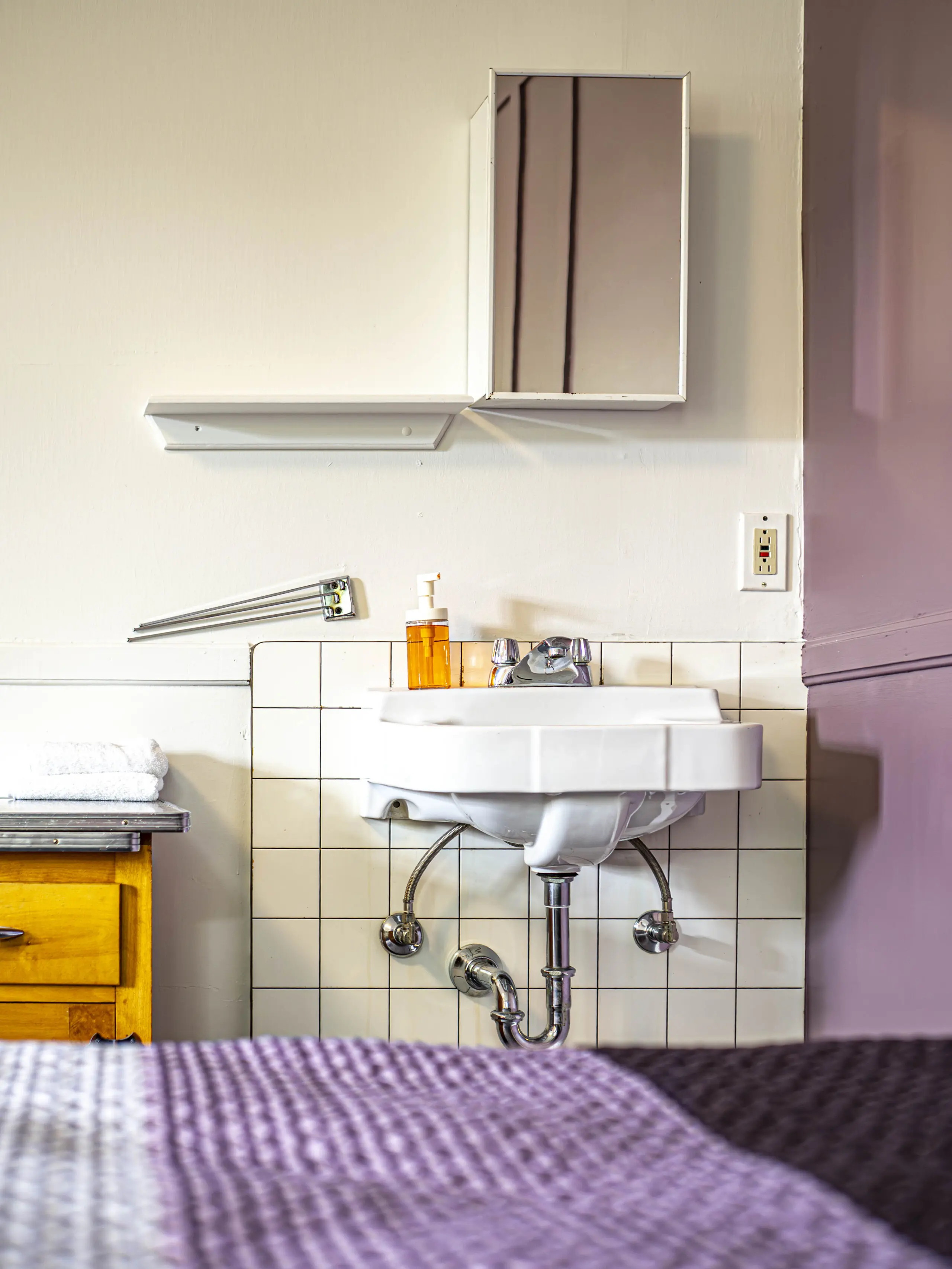 Un petit lavabo mural avec un distributeur de savon au-dessus de carreaux blancs, une armoire de toilette, une serviette pliée sur un comptoir en bois, et une partie d'un couvre-lit violet au premier plan.
