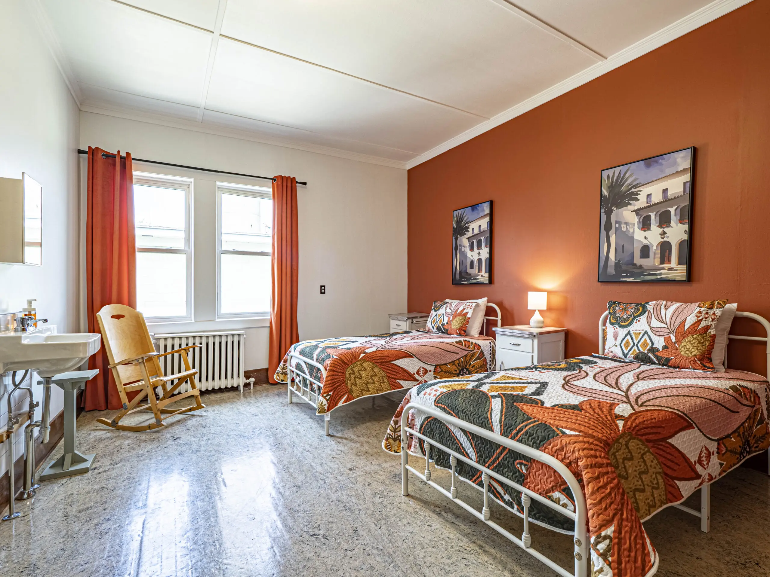 Une chambre lumineuse avec deux lits jumeaux en métal avec une literie à fleurs orange, un mur d'accent orange, deux tableaux encadrés, une table de nuit avec une lampe, une chaise à bascule et de grandes fenêtres avec des rideaux orange.