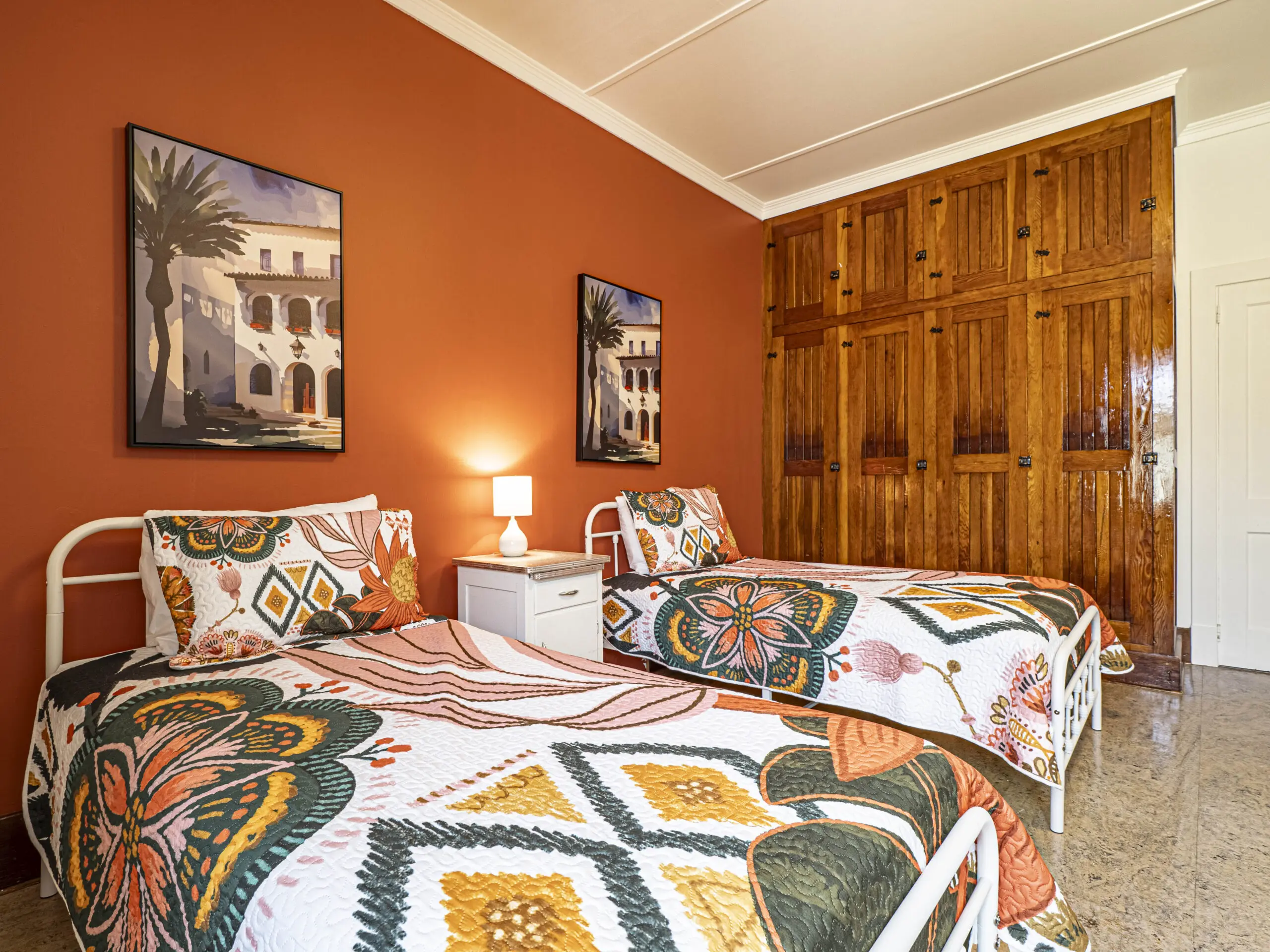 Une chambre lumineuse avec deux lits simples dotés d'une literie colorée à motifs, une table de nuit blanche avec une petite lampe, des murs orange, des armoires encastrées en bois et des tableaux encadrés représentant des bâtiments et des palmiers.