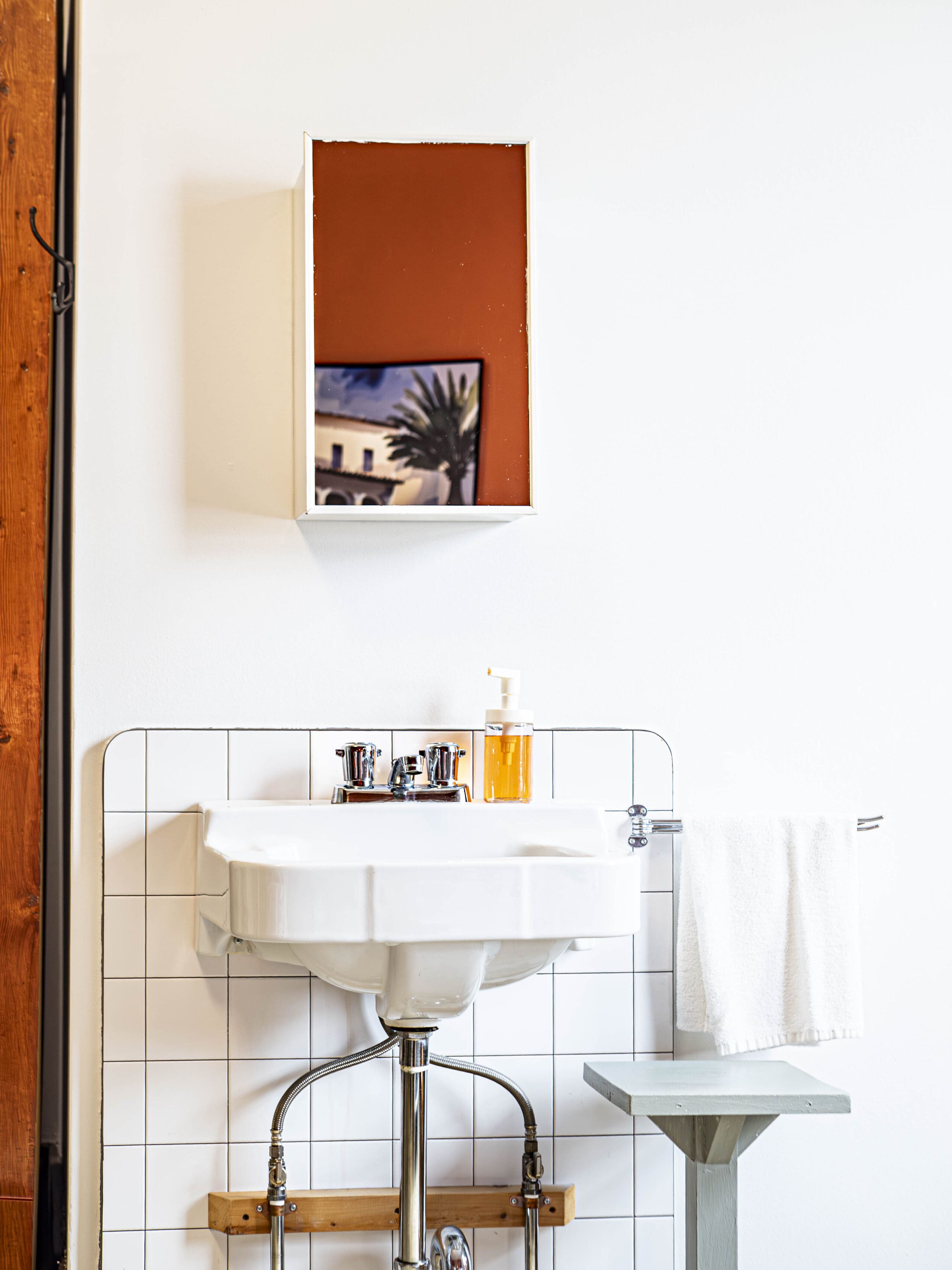 Un évier blanc avec des accessoires chromés est fixé à un mur carrelé. Au-dessus, une étagère contient une photo encadrée et un distributeur de savon. Une serviette blanche est suspendue à un support et une petite table se trouve à proximité, contre un mur blanc.