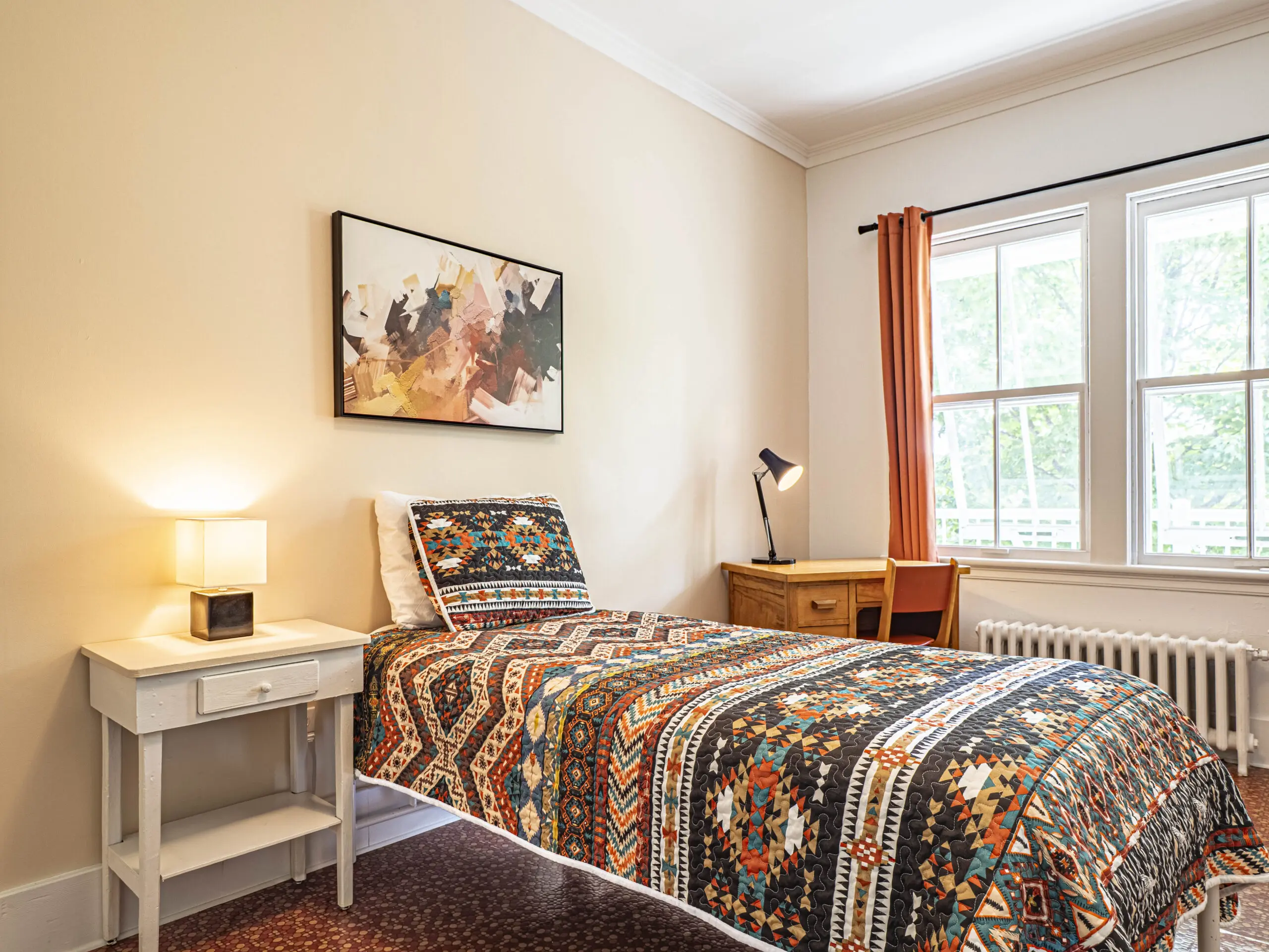 Une chambre à coucher confortable avec un lit simple recouvert d'une courtepointe à motifs géométriques colorés, une table de nuit blanche avec une lampe, un bureau en bois avec une lampe noire, et une fenêtre aux rideaux orange laissant entrer la lumière naturelle.