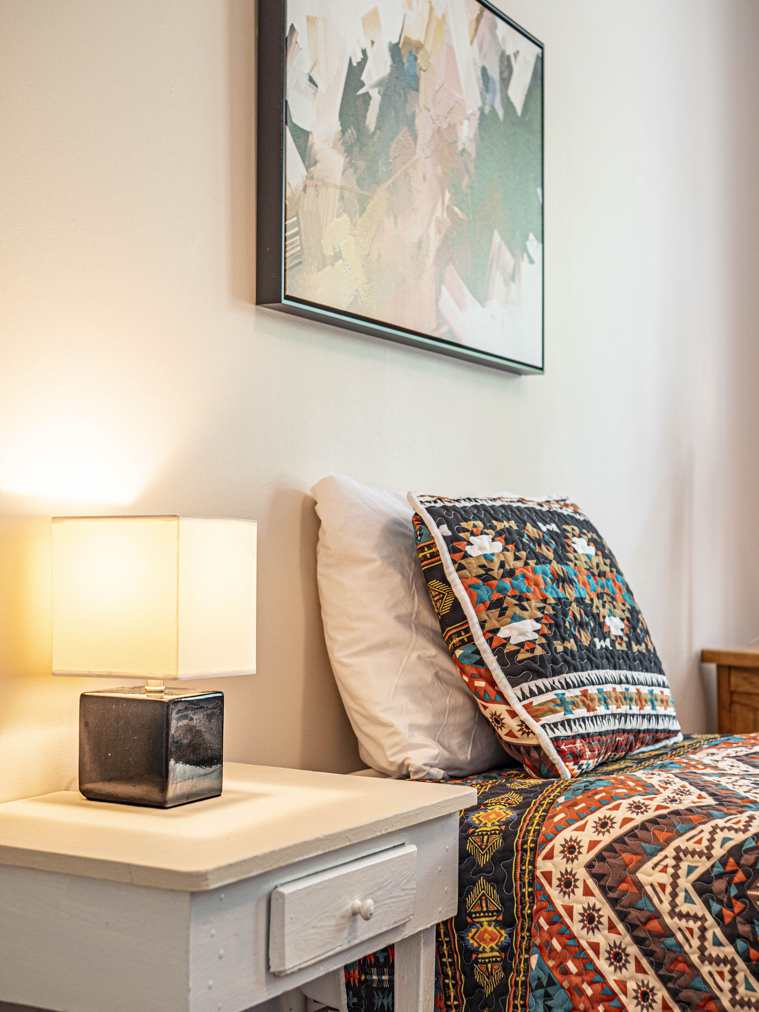 Une table de chevet avec une lampe carrée se trouve à côté d'un lit recouvert d'un édredon coloré à motifs géométriques, avec une peinture abstraite accrochée au mur au-dessus du lit.