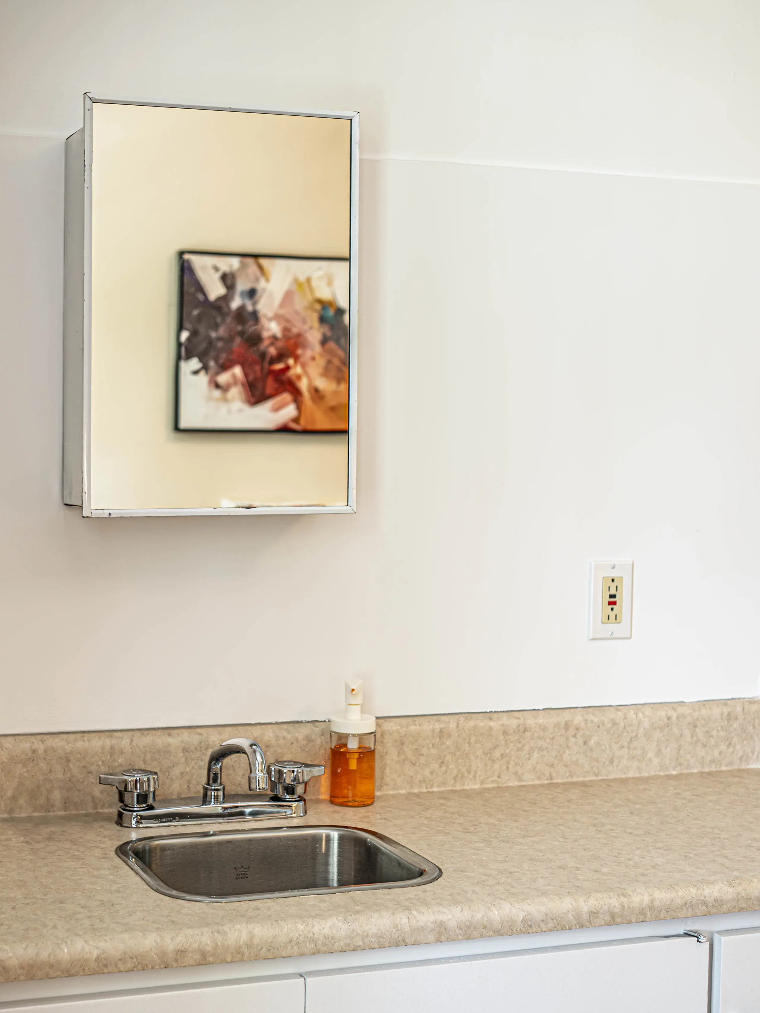 Un petit évier en acier inoxydable avec un distributeur de savon se trouve sous une armoire à pharmacie en miroir, qui reflète une œuvre d'art abstraite floue. Le plan de travail est beige et une prise électrique est visible sur le mur.