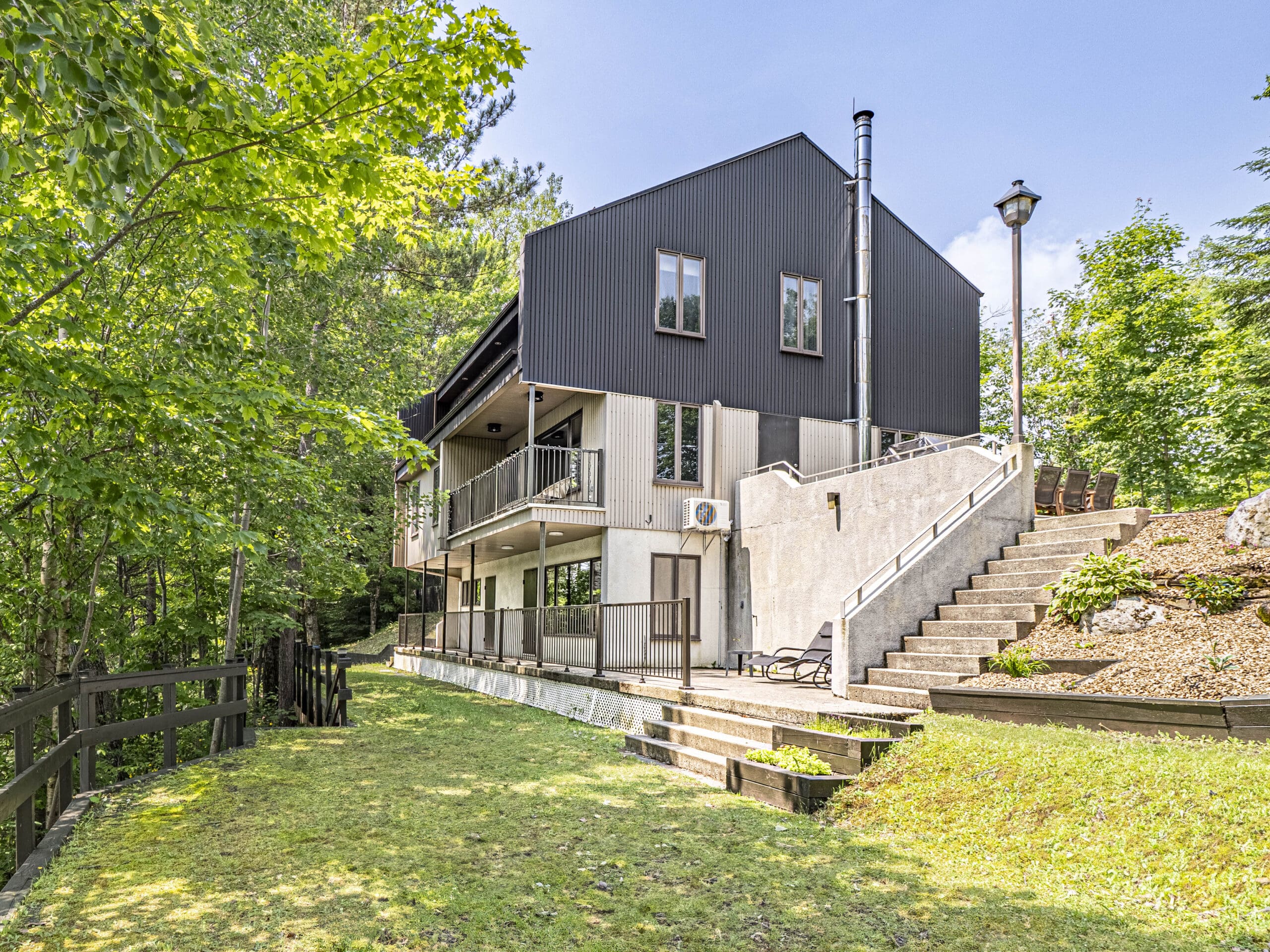 Maison moderne à deux étages, à l'extérieur noir et blanc, entourée d'arbres verdoyants. La maison est dotée de grandes fenêtres, de balcons et d'un patio en gradins avec des chaises longues. Une cour clôturée et un escalier extérieur complètent la scène.